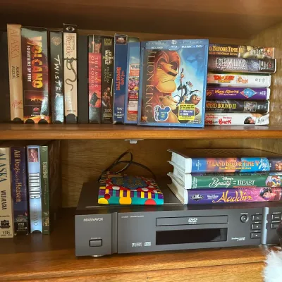 Sale Photo Thumbnail #194: Magnavox DVD/CD player displayed on wooden shelves alongside a collection of VHS tapes and a copy of Disney's The Lion King on Blu-ray.