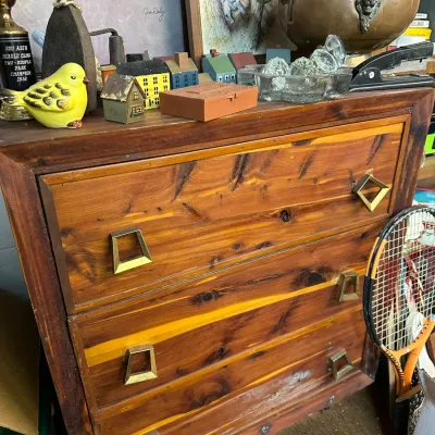 Sale Photo Thumbnail #54: Vintage mid-century knotty pine or cedar three-drawer chest featuring distinct trapezoidal metal handles, topped with various collectibles including framed prints, a ceramic bird figurine, and a brass coal scuttle.