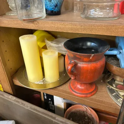 Sale Photo Thumbnail #290: Rustic red urn-style ceramic planter sitting alongside pillar candles on a brass plate and miscellaneous glassware displayed on wooden shelving.