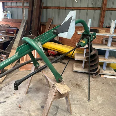 Sale Photo Thumbnail #753: John Deere green 3-point hitch post hole digger with PTO shaft, resting on wooden sawhorses in a workshop.
