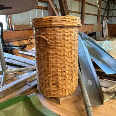 Sale Photo Thumbnail #535: Tall, cylindrical woven wicker hamper with a matching lid and side handles, sitting in a barn surrounded by miscellaneous items.