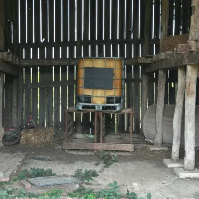 Sale Photo Thumbnail #756: Large caged IBC tote water tank elevated on a wooden stand inside a rustic barn structure.
