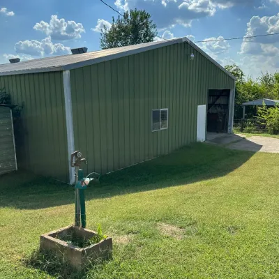 Sale Photo Thumbnail #276: Large green metal pole barn building featuring an open garage door and a outdoor water spigot setup in the foreground lawn.