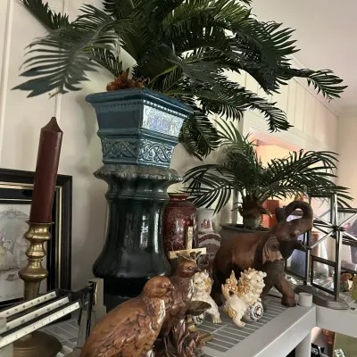 Sale Photo Thumbnail #660: Collection of decorative home accents, including a tall glazed ceramic pedestal with an artificial palm, ceramic bird figurines, Foo dogs, and a carved wooden elephant statue.