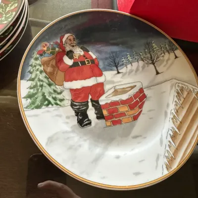 Sale Photo Thumbnail #233: Decorative holiday plate featuring an illustration of Santa Claus with a toy sack beside a rooftop chimney, accented with a gold rim.