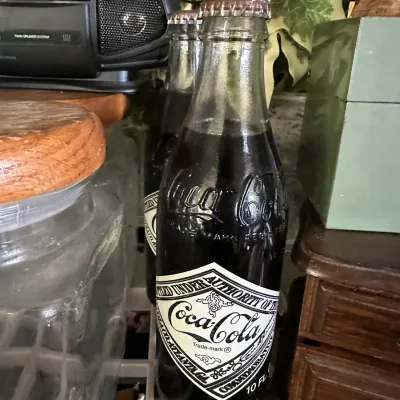 Sale Photo Thumbnail #236: Vintage filled commemorative 10 fl oz Coca-Cola glass bottles with original metal crown caps and diamond paper labels.