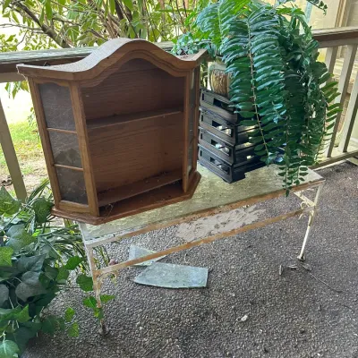 Sale Photo Thumbnail #183: Vintage small wooden wall curio cabinet with glass side panels resting on a wrought iron plant bench alongside potted greenery.