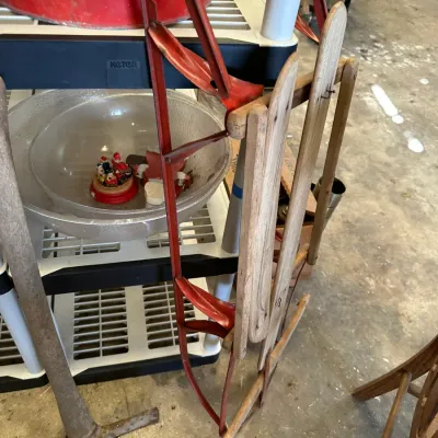 Sale Photo Thumbnail #529: Vintage wooden sled with painted red metal runners resting against a plastic storage shelf next to a pickaxe.
