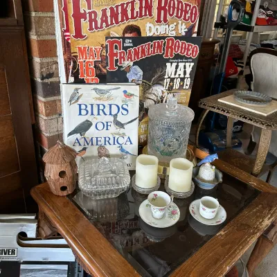 Sale Photo Thumbnail #642: Collection of decorative tabletop items including Franklin Rodeo posters, a 'Birds of America' book, cut glass jars, LED pillar candles, and miniature teacups displayed on a wooden end table.