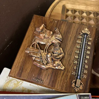 Sale Photo Thumbnail #117: Vintage Alaska souvenir wall thermometer mounted on faux-wood plaque, featuring a raised copper-tone gold prospector and pack mule emblem.