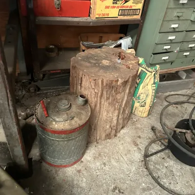 Sale Photo Thumbnail #747: Vintage galvanized metal gas can with red trim sitting beside a large wood chopping stump in a workshop, surrounded by storage shelves and tools.