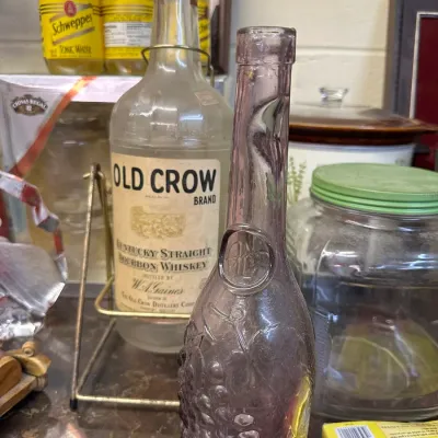 Sale Photo Thumbnail #34: Vintage amethyst glass bottle with embossed grape design alongside a large Old Crow bourbon glass jug in a metal caddy.