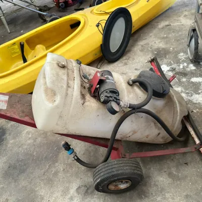 Sale Photo Thumbnail #543: Portable pull-behind chemical or lawn sprayer featuring a poly tank mounted on a wheeled metal frame, complete with 12V pump and pressure gauge. Shows heavy dirt and signs of outdoor use.