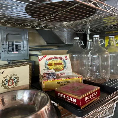 Sale Photo Thumbnail #80: Collection of vintage cigar boxes, including Don Diego, Tampa Nugget, and Roi-Tan, alongside clear glass gallon jug bottles stored on wire shelving.