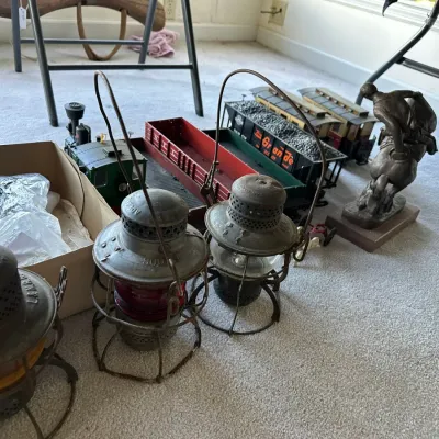 Sale Photo Thumbnail #198: Collection of vintage railroad lanterns with colored glass globes displayed alongside model train cars and a bronze cowboy statue on a carpeted floor.