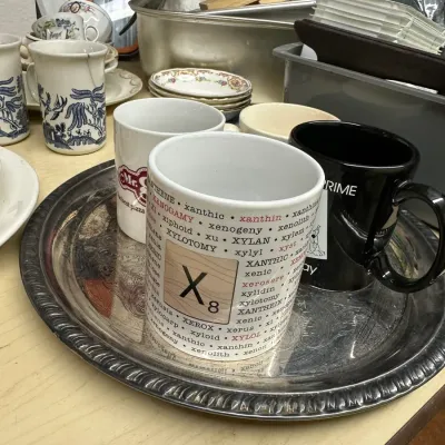 Sale Photo Thumbnail #6: Assorted ceramic coffee mugs displayed on an engraved silver-plated tray, including a Scrabble-themed 'X' mug and a Blue Willow patterned mug.