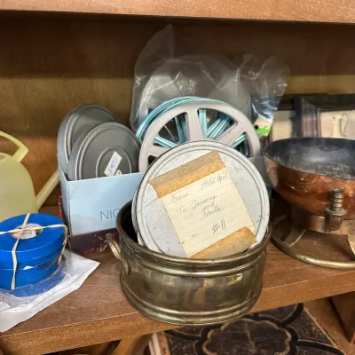 Sale Photo Thumbnail #64: Collection of vintage home movie reels and metal film canisters, including one labeled "Cruise 1972 April To Jamaica and Haiti," sitting on a wooden shelf.