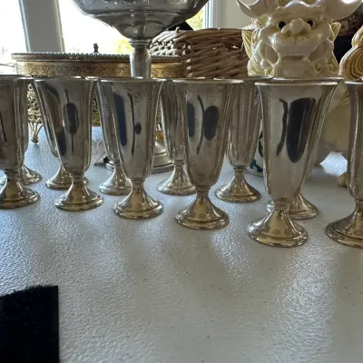 Sale Photo Thumbnail #389: Collection of vintage silver-plated cordial stems displaying patina, arranged alongside decorative porcelain pieces on a table.
