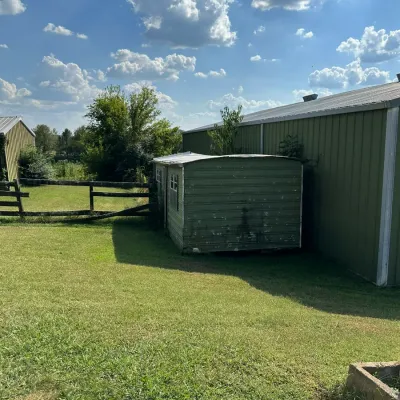 Sale Photo Thumbnail #277: Weathered green metal storage shed situated next to a larger barn on a grassy farm property.