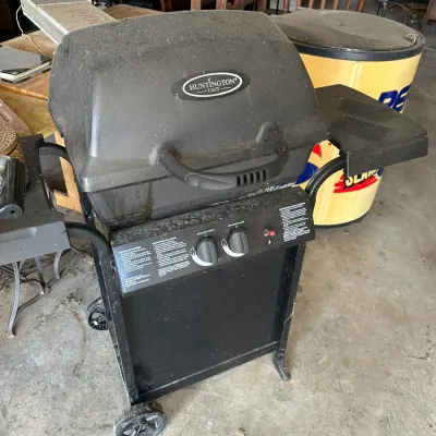 Sale Photo Thumbnail #711: Huntington Cast two-burner propane gas grill on a wheeled black metal cart with side shelves.