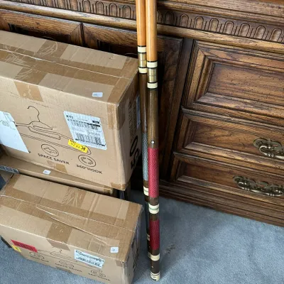 Sale Photo Thumbnail #379: Pair of vintage two-piece wooden pool cues with decorative thread wraps and joint rings, leaning against a carved wooden cabinet.