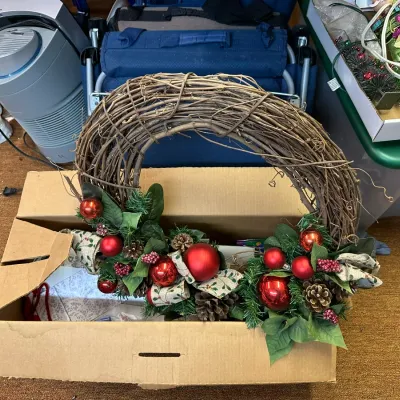 Sale Photo Thumbnail #315: Rustic grapevine Christmas wreath decorated with red ornaments, faux greenery, pinecones, and ribbon, packed in a cardboard box alongside other household items.