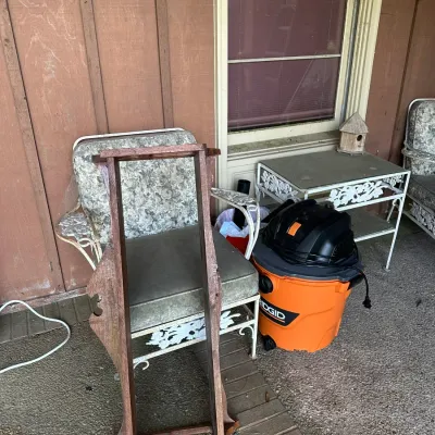 Sale Photo Thumbnail #186: A wooden gun or fishing rod rack leaning against a vintage wrought iron patio armchair, alongside an orange RIDGID wet/dry shop vacuum on a porch.
