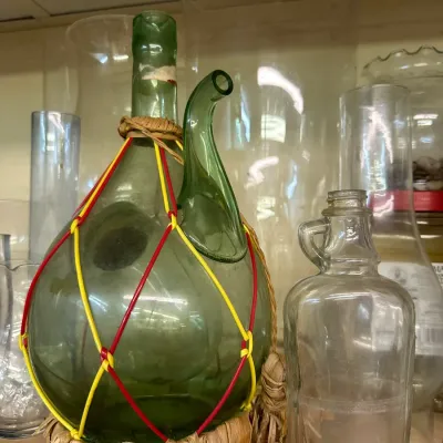 Sale Photo Thumbnail #61: Vintage green glass porron wine decanter encased in a woven raffia basket with colorful string netting, displayed alongside clear glassware.