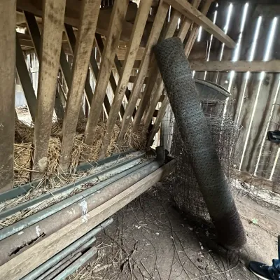 Sale Photo Thumbnail #339: A roll of dark wire fencing and wire mesh stored inside a rustic barn next to a wooden hay feeder with metal troughs.
