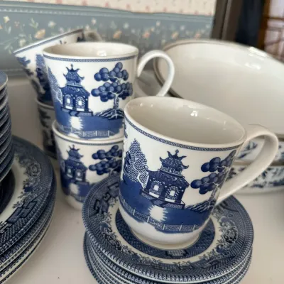 Sale Photo Thumbnail #696: Vintage Blue Willow pattern porcelain tableware, featuring coffee mugs, stacked saucers, and plates in classic blue and white Asian-inspired pagoda design.