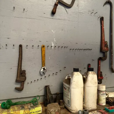 Sale Photo Thumbnail #479: Assorted vintage hand tools, including pipe wrenches, a hacksaw, adjustable wrench, and pry bar, hanging on a workshop pegboard alongside workbench supplies.