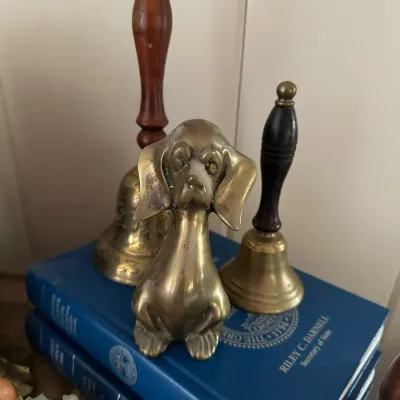 Sale Photo Thumbnail #245: Vintage brass dog figurine displayed with two wooden-handled brass hand bells resting atop a stack of blue hardcover books.