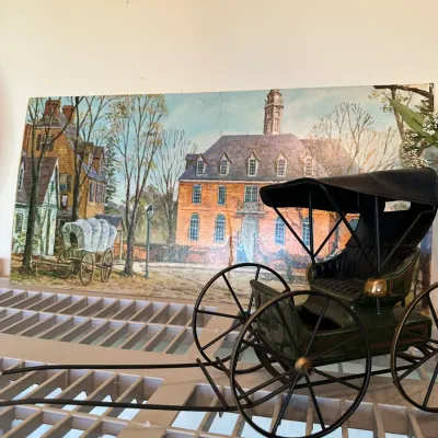 Sale Photo Thumbnail #674: Miniature horse-drawn buggy model with spoked metal wheels and a vinyl canopy, displayed in front of a colonial Williamsburg scene art print.
