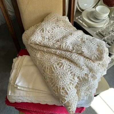 Sale Photo Thumbnail #662: Stack of vintage linens featuring a large handmade cream crochet tablecloth, scalloped white cloth napkins, and bright pink ribbed placemats.