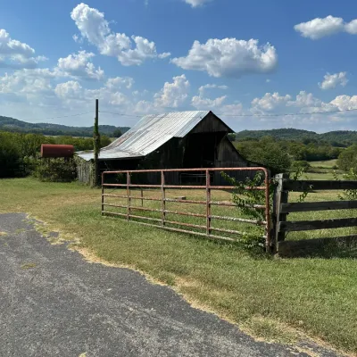 Sale Photo Thumbnail #472: Rustic weathered wooden barn with a metal roof set behind a red metal livestock gate in a rural landscape.