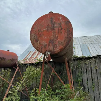 Sale Photo Thumbnail #341: Vintage elevated red metal fuel tank on a steel stand, showing heavy rust and weathering from outdoor use.