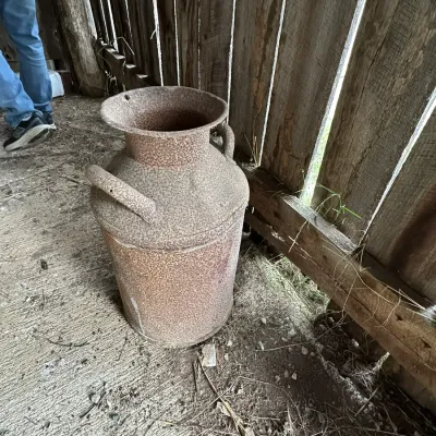 Sale Photo Thumbnail #329: Vintage metal milk can with heavy surface rust and side handles, sitting outdoors against a wooden fence.