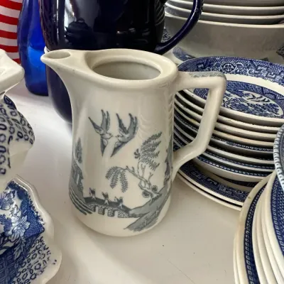 Sale Photo Thumbnail #701: Vintage white pottery creamer pitcher with a grey Blue Willow pattern transferware design, displayed alongside stacks of matching blue-and-white dishware.