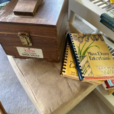 Sale Photo Thumbnail #263: Vintage Kiwi Hand Crafted wooden shoe valet box alongside spiral-bound cookbooks, resting on a tan upholstered footstool.