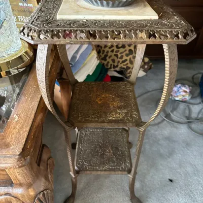 Sale Photo Thumbnail #685: Vintage brass multi-tiered plant stand featuring elaborate embossed floral patterns, cabriole legs, and a marble top insert with a small fluted metal dish.