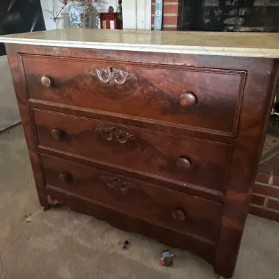 Sale Photo Thumbnail #584: Antique marble-top three-drawer dresser featuring carved wood accents, flame mahogany veneer, and wooden knobs, showing some wear along the bottom edge.