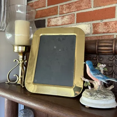 Sale Photo Thumbnail #257: Vintage polished brass picture frame with rounded corners alongside a glass hurricane candle holder and a porcelain bluebird figurine.