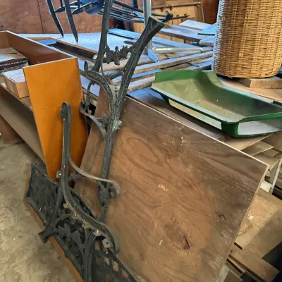 Sale Photo Thumbnail #534: Pair of ornate cast iron garden bench side frames leaning against wooden boards, alongside miscellaneous items including a wicker hamper and plastic tray.