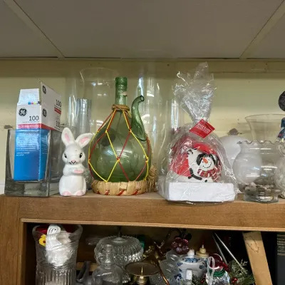 Sale Photo Thumbnail #296: Shelf displaying assorted glassware and holiday decor, including a vintage wicker-wrapped green glass porron, ceramic bunny figurine, snowman decorations, and glass vases.