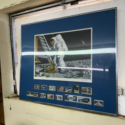 Sale Photo Thumbnail #33: Framed Apollo moon landing print featuring a central artwork of an astronaut on the lunar surface above a collection of space-themed postage stamps, set in a blue mat and silver-tone frame.