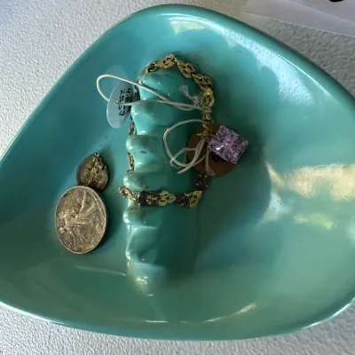 Sale Photo Thumbnail #408: Mid-century turquoise ceramic triangle ashtray filled with a gold chain bracelet featuring purple gemstone accents, a pendant, and vintage coins.
