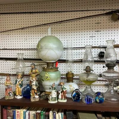 Sale Photo Thumbnail #45: Collection of vintage oil lamps, porcelain figurines, and art glass paperweights displayed on a shelf in front of pegboard-mounted fishing rods.