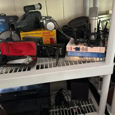 Sale Photo Thumbnail #659: White plastic shelving unit holding vintage cameras, electronics, studio microphone with pop filter, and hardback books.
