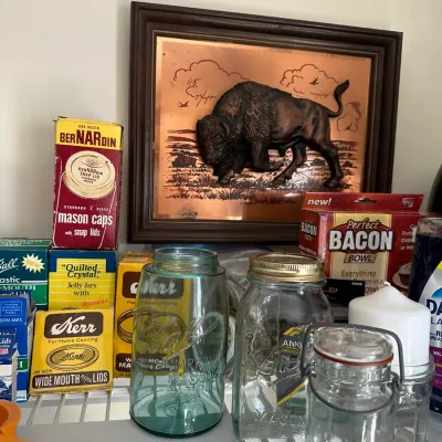 Sale Photo Thumbnail #664: A collection of vintage glass canning jars, including a blue Ball Perfect Mason jar, stacked boxes of lids and supplies, and a framed copper relief plaque featuring an American bison.