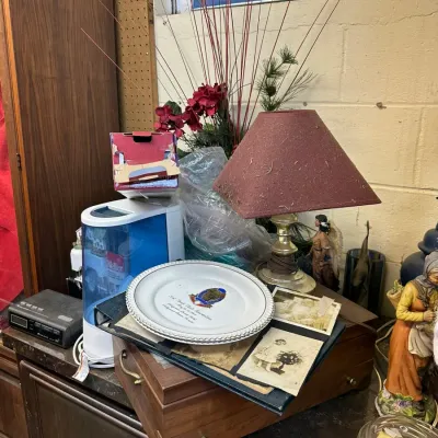 Sale Photo Thumbnail #314: Assorted household tabletop goods including a small brass table lamp with a maroon shade, a commemorative plate resting on a photo album, a desktop humidifer, and decorative figurines.